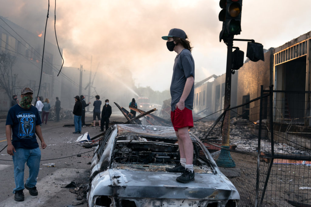 A man stands on a burned out car on Thursday morning as fires burn behind him in the Lake St area of Minneapolis, Minnesota. Photo: Lorie Shaull from St Paul, United States